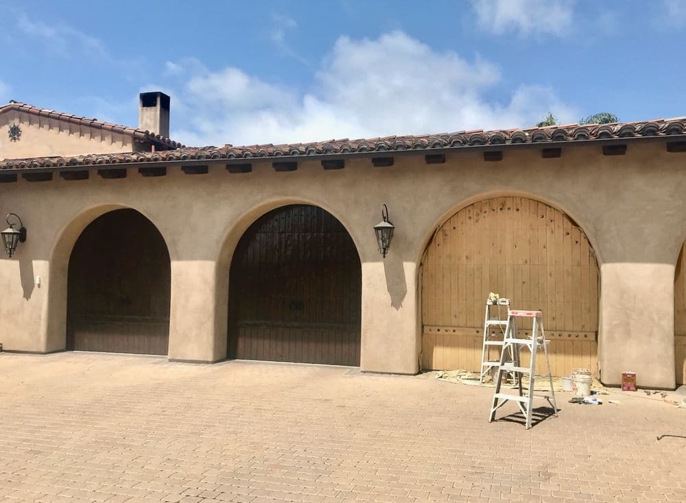 Arched garage doors with wooden panels and a ladder in front against a cloudy sky.