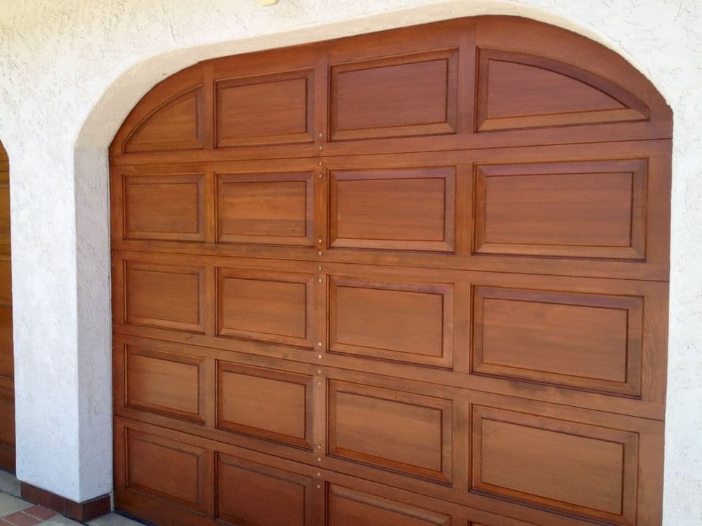 Wooden garage door with decorative panels and arched top against a stucco wall.