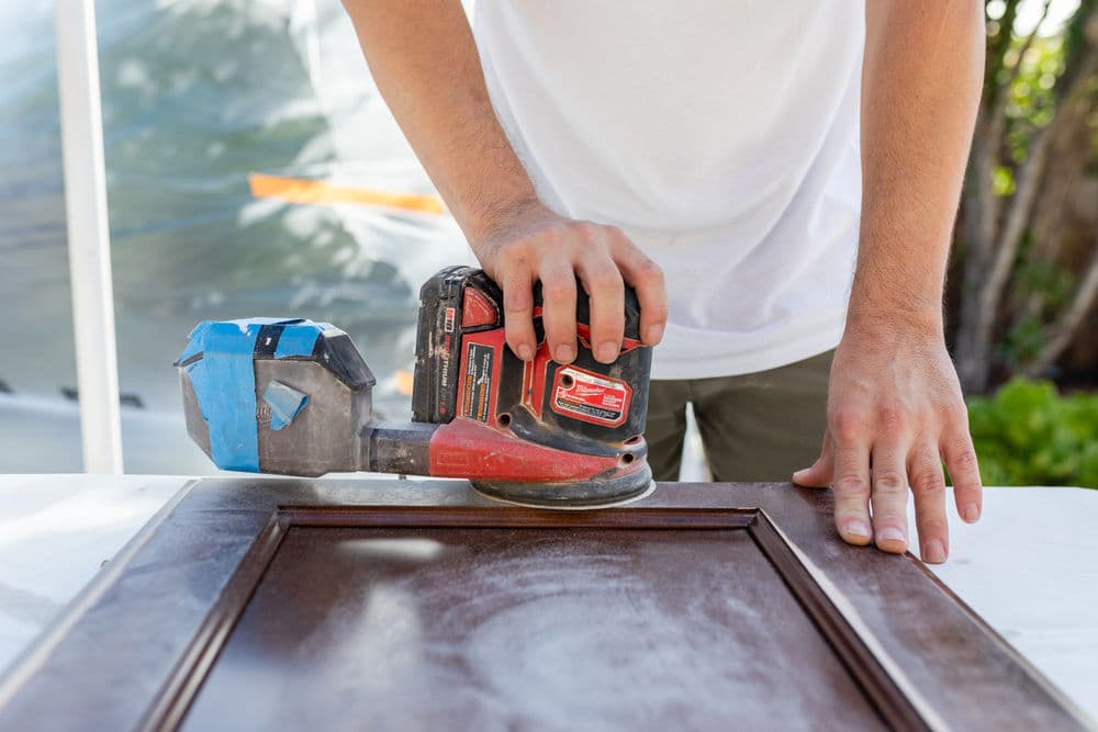 Person using a power sander on a wood surface in a bright outdoor workspace.